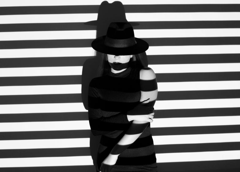 Black and white of anonymous female in hat standing near wall with striped pattern against projector with shadow on face
